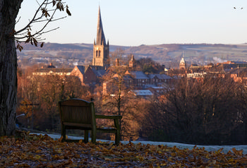 Tapton park winter spire close This landscape photograph was taken in the morning during winter at Tapton Park in Chesterfield, United Kingdom. The main subject of the image is the iconic spire of the Church of St Mary and All Saints, which is clearly visible rising above the town’s buildings. The church spire stands out against the backdrop of rolling hills and the frosted ground, highlighting the winter atmosphere. Nature is prominent in the foreground, with fallen leaves covering the frosty park floor, bare trees framing the scene, and a wooden bench positioned under a large tree. Tapton Park provides an elevated view over Chesterfield, with trees and the town’s architecture taking centre stage. This view is closely associated with the famous crooked spire, a notable landmark in Chesterfield and the wider region of the United Kingdom.